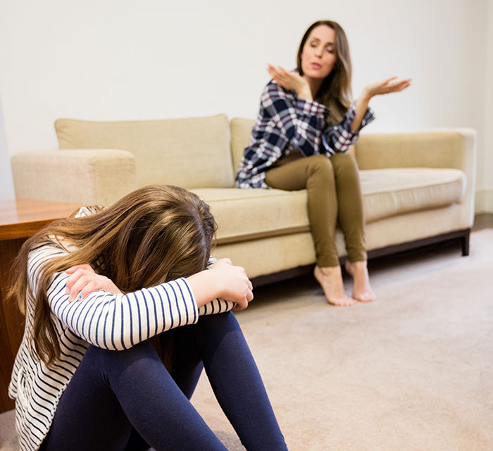 Woman frustrated sitting on couch arguing with upset daughter sitting on floor hugging knees