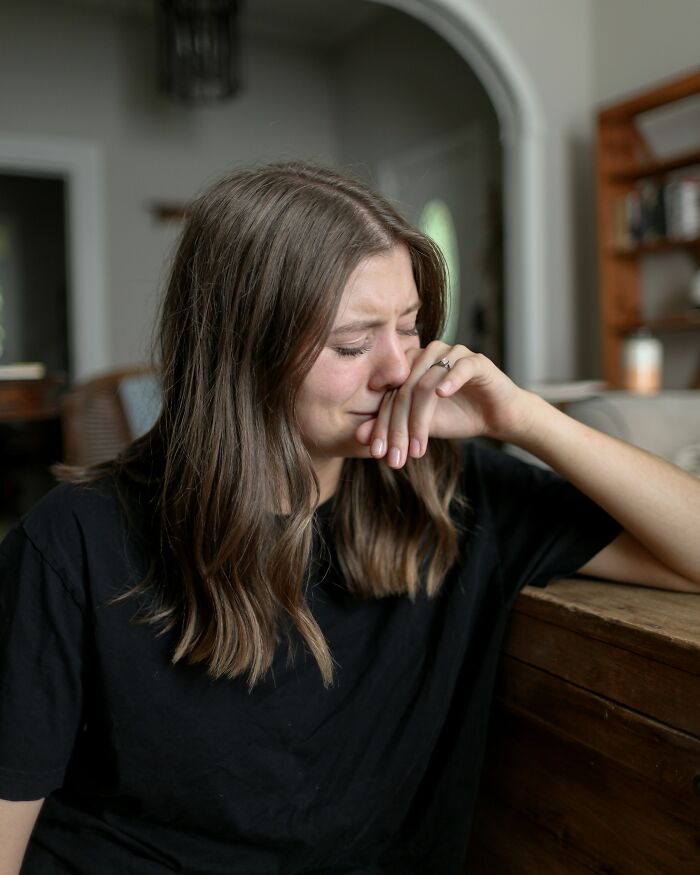 A young woman with long brown hair, wearing a black shirt, covering her mouth while crying, reflecting on deeply immoral things.