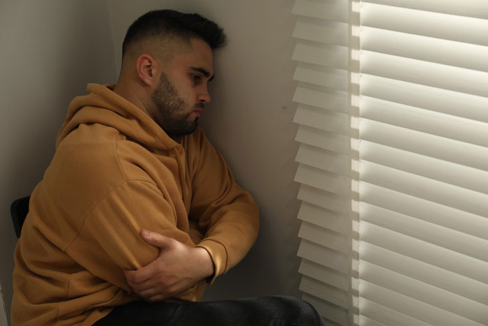 Man in a hoodie, head bowed, leaning against a wall by a window, possibly stressed about brother wedding money food.