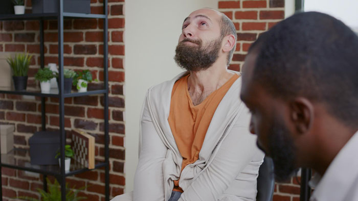 A man, eyes looking up in distress, sits beside another man, discussing issues like wedding money or food.