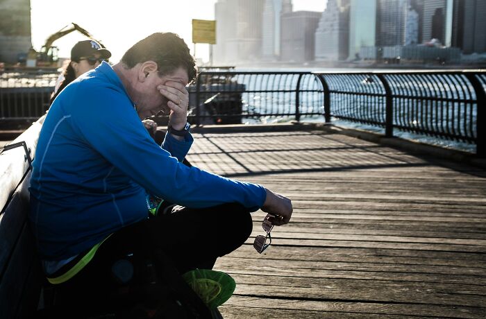 Man on a pier, distressed, holding sunglasses, contemplating health warning signs or Routine Cancer Screening. City skyline in background.