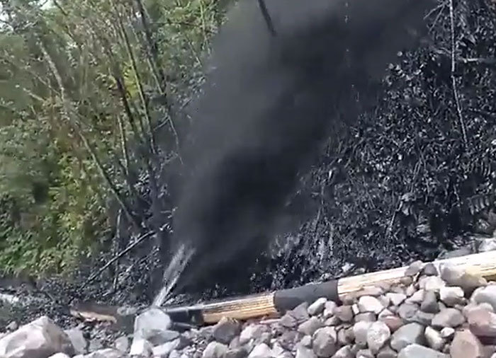 A pipe leaks catastrophically, spewing a dark liquid onto a rocky riverbank, staining the nearby foliage black.