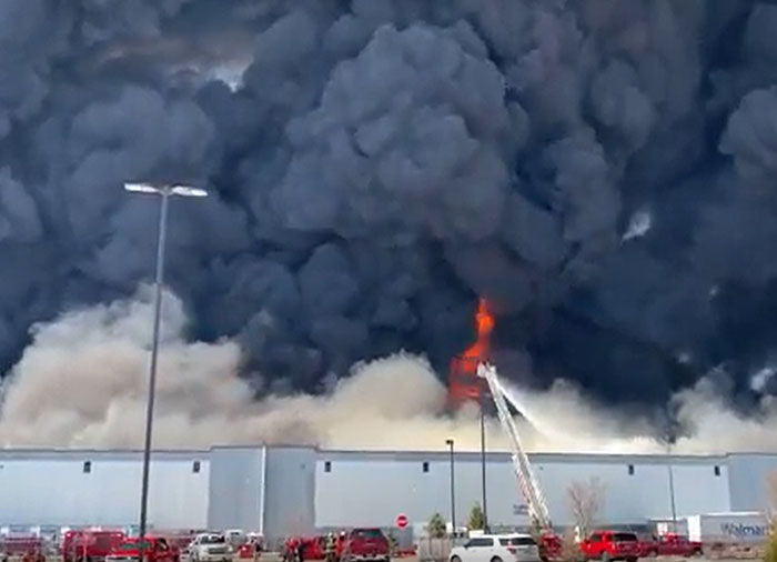 Massive fire with black smoke billowing from a machinery or structure, firefighters attempting to extinguish the catastrophically failed building.