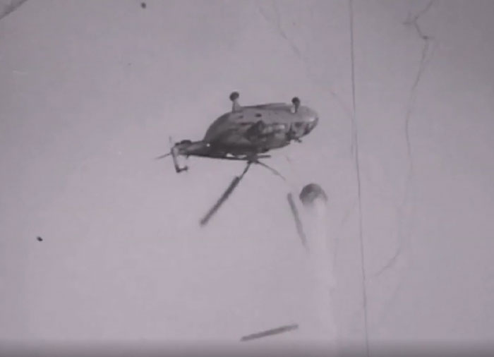 A black and white image of a helicopter failing catastrophically, with parts detaching and falling from the sky.
