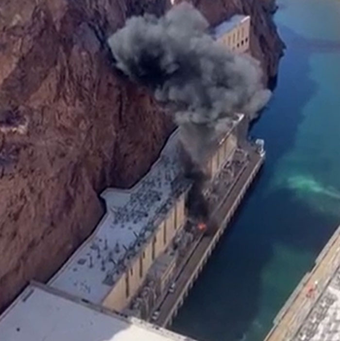 Aerial view of a structure with thick black smoke rising, showing catastrophic machinery failure next to a body of water.