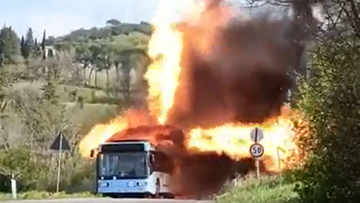A white bus engulfed in flames with huge plumes of fire and smoke billowing, a catastrophic machinery failure.