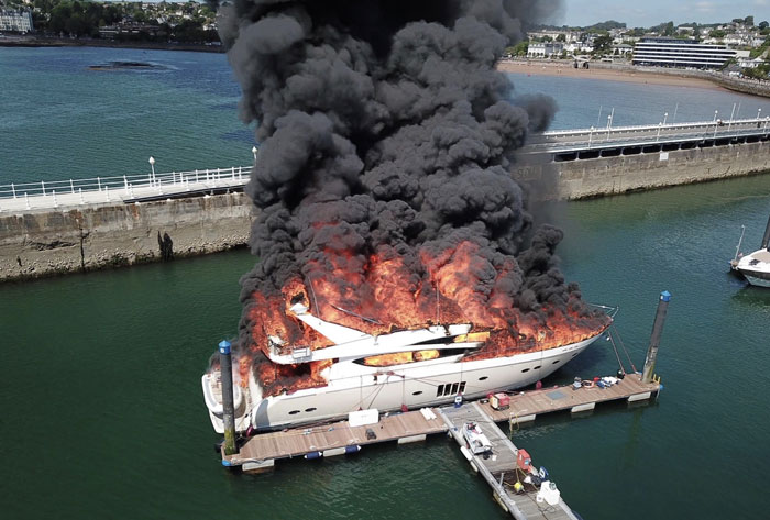 A large white yacht engulfed in flames and thick black smoke, showing machinery that has failed catastrophically in a harbor.