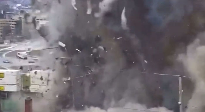 A structure catastrophically failed in an urban setting, enveloped by a large plume of smoke and debris mid-explosion.