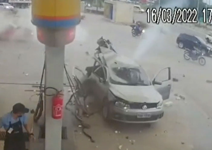 CCTV captures a grey car catastrophically failed after crashing into a gas station pump, with debris and a worker reacting.