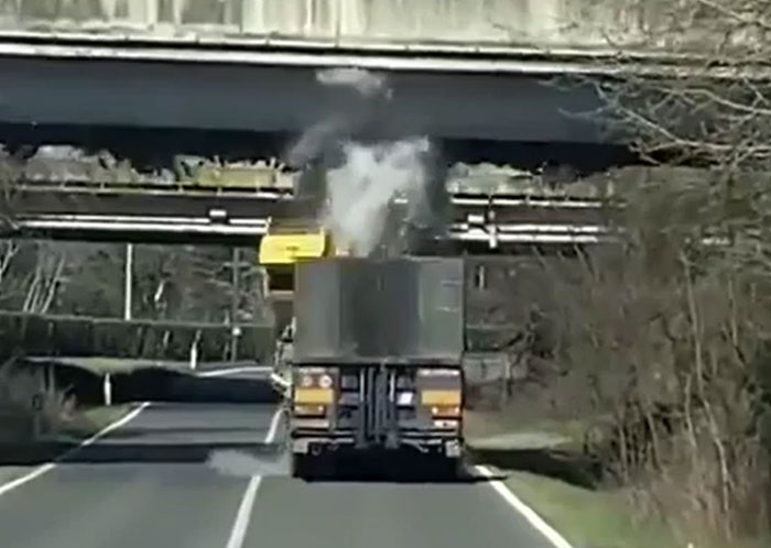 A large truck with a raised bed drives under an overpass, generating a large puff of white smoke from machinery that has failed catastrophically.