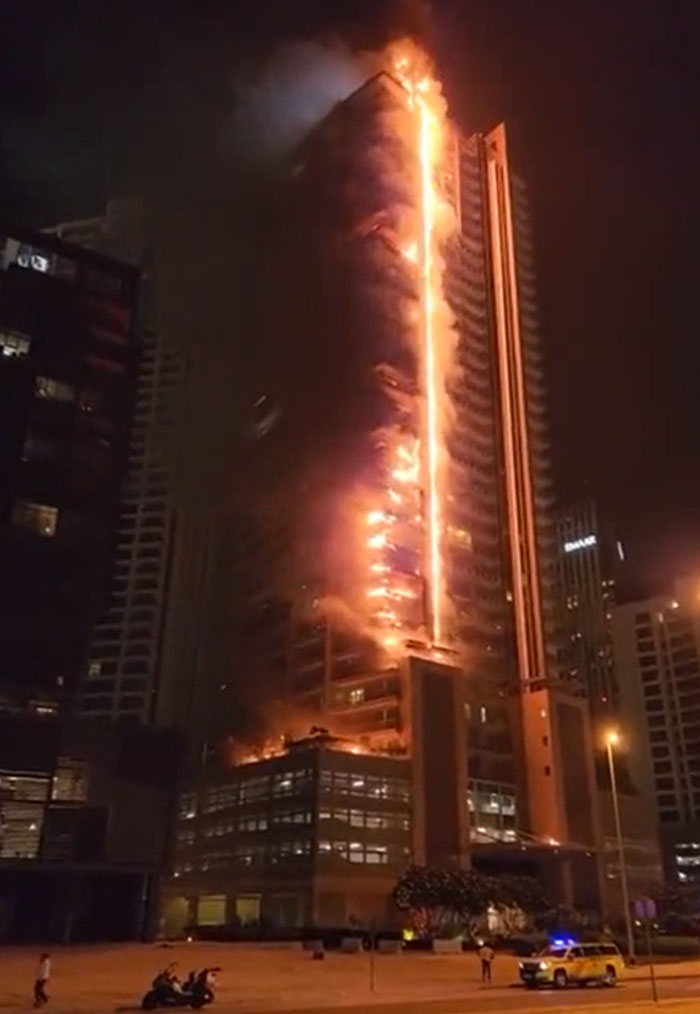 Nighttime view of a tall skyscraper engulfed in a catastrophic fire, flames and smoke visible along its entire height.