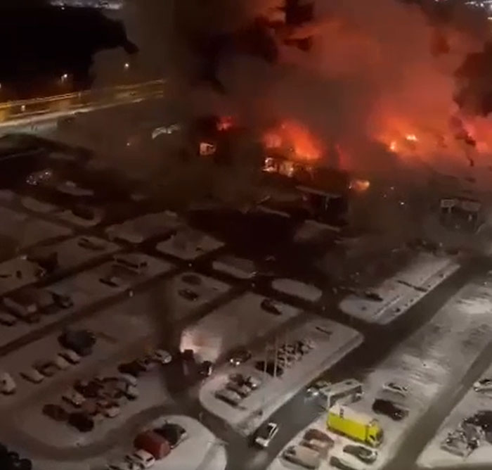 Aerial night view of a large building engulfed in flames, a catastrophic machinery failure, with a snow-covered parking lot below.