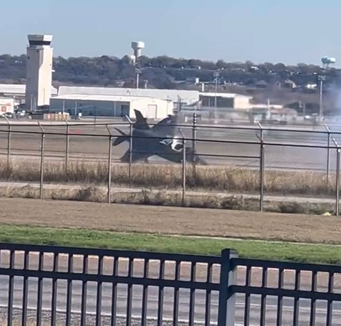 A military jet on a runway, nose-down with smoke, showing a machinery failure, behind a fence.