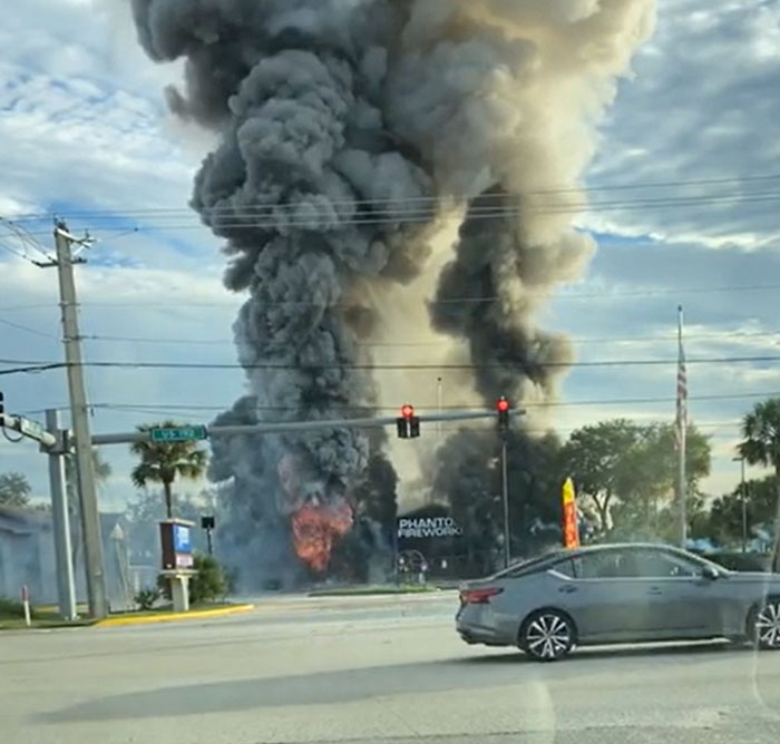Catastrophically failed fireworks store engulfed in fire, emitting massive black smoke into the sky.