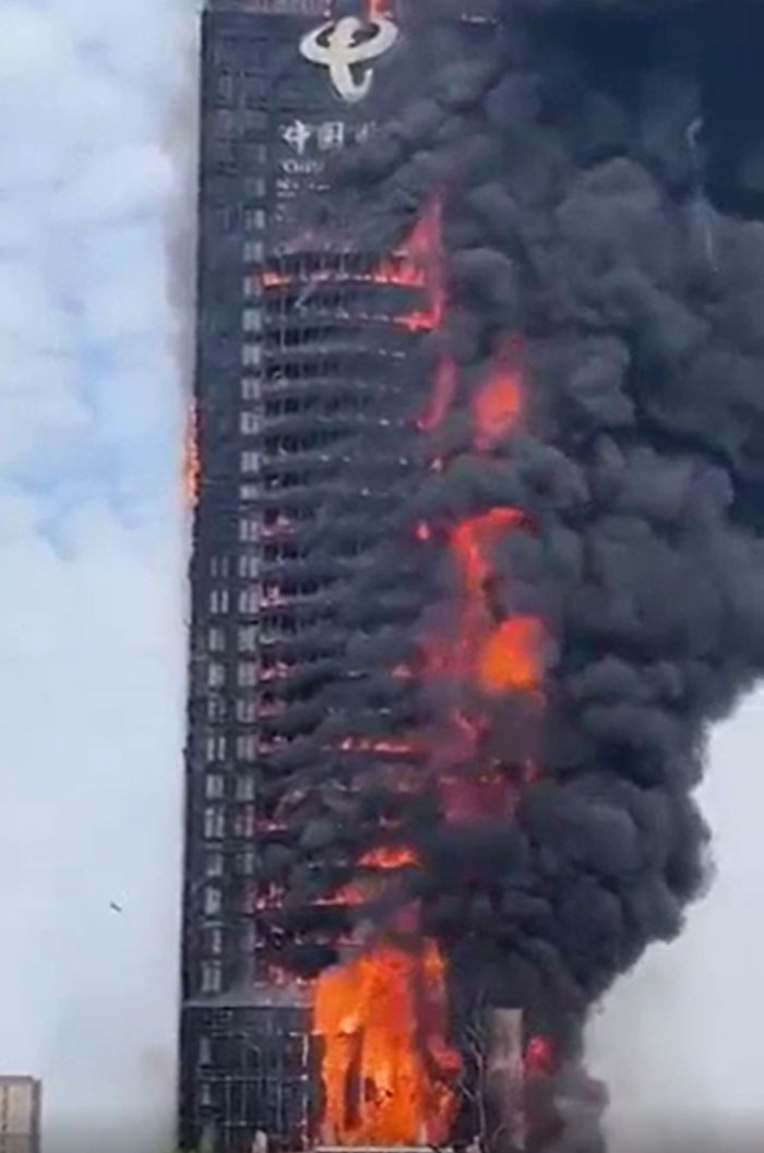 A tall building engulfed in flames and dense black smoke, a catastrophic failure. Bright orange fire erupts from many windows.