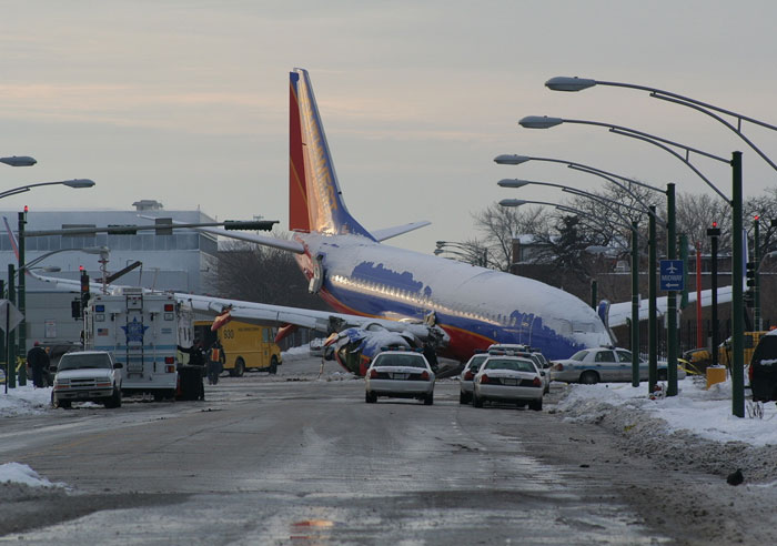 A snow-covered airplane, a machinery failure, on a street with police cars and emergency vehicles after a catastrophic accident.