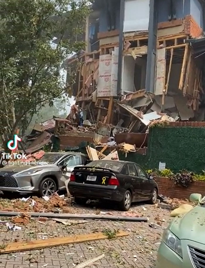 A house structure that has failed catastrophically, showing extensive damage and debris, with cars parked nearby.