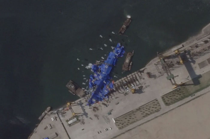 An aerial view of a large blue structure, possibly machinery, catastrophically failed in a harbor next to a dock.