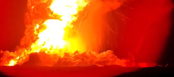 A massive, catastrophic fire engulfs the night sky with brilliant orange and red flames, showing machinery failure.