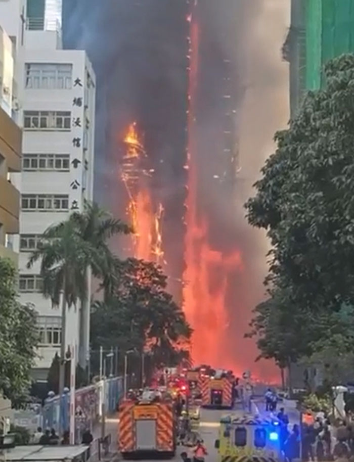 A towering skyscraper engulfed in massive flames, with fire engines and emergency vehicles on the street below; a machinery failure catastrophe.