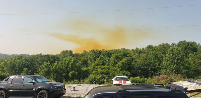 A large plume of orange-brown smoke rises from machinery that has failed catastrophically over a green forest.