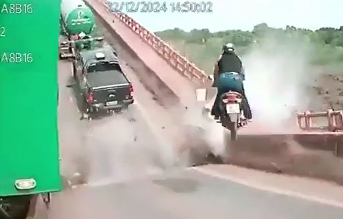 A white motorcycle passes a truck towing a trailer with a tank, amidst machinery failure and smoke.
