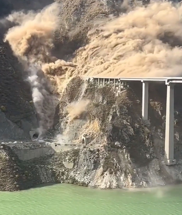 Massive landslide engulfing a bridge and road, demonstrating structures that have failed catastrophically. Mud and dust cascade into green water.
