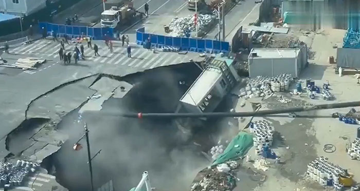 An aerial view of a huge sinkhole on a street, with a building partially collapsed into it, showing machinery and structures failed catastrophically.