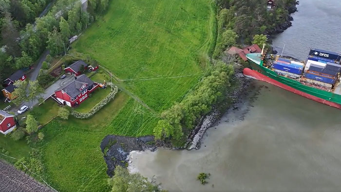 Aerial view of a cargo ship that failed catastrophically, running aground near houses and green fields by the water.