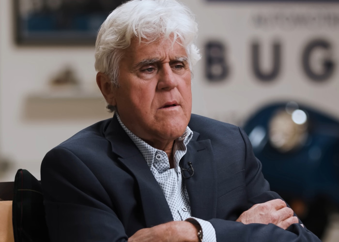 Jay Leno, with his signature white hair, wearing a dark suit and patterned shirt, exemplifies frugal celebrity saving habits.