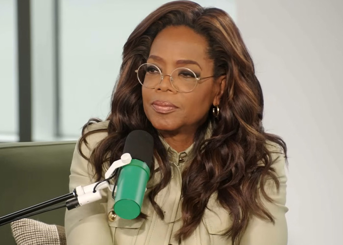 Oprah Winfrey, wearing glasses and a light jacket, speaking into a microphone about how to be frugal.