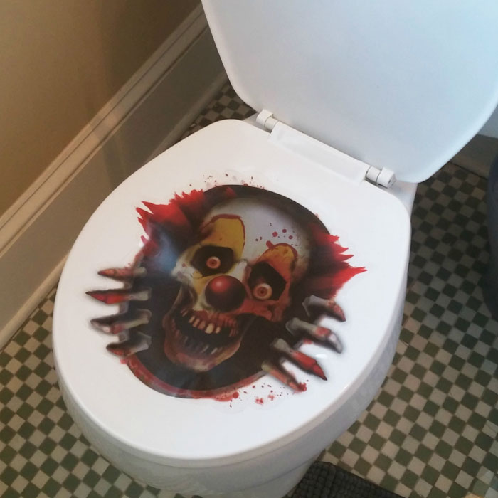 A toilet seat with a terrifying clown decal, showing a skull-like clown with red hair and bloody hands.