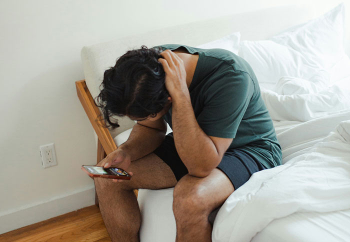 A man sits on a bed, head in his hand, looking at his phone, realizing his fianc&eacute;e left him for cheating.