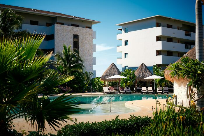 Hotel pool area with loungers and umbrellas surrounded by tropical plants and buildings