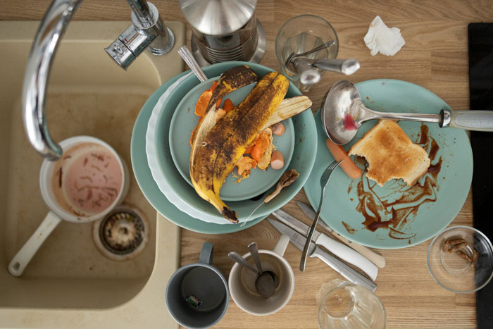 An overhead shot of dirty dishes, cutlery, food scraps, and a banana peel piled in a sink and on a countertop. This image highlights the issue of a woman expected to be a maid.