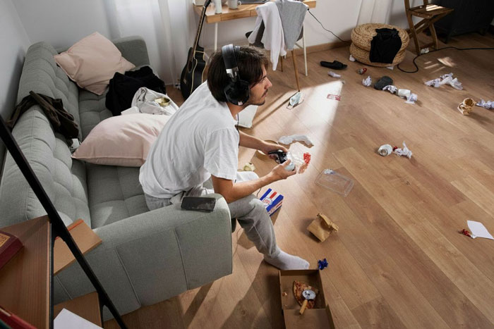 A man with headphones plays video games on a messy floor with trash, highlighting a woman's "maid" dilemma.