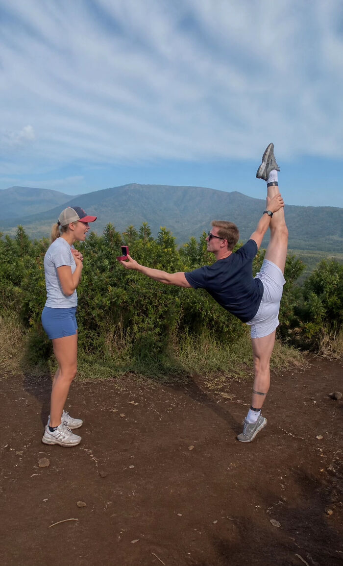 Man doing a high kick while proposing to a woman, showing a proposal pic in a scenic mountain setting.