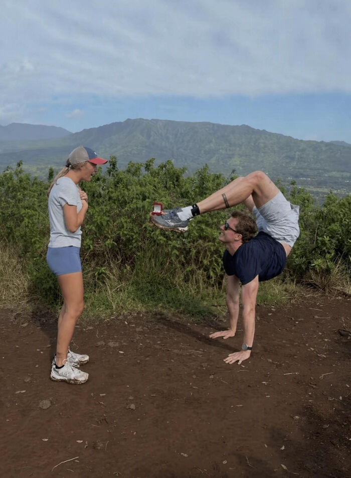 A man in a handstand doing a proposal pic, holding a ring box on his foot for a surprised woman.