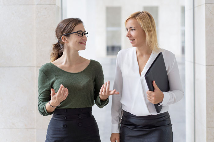 Two women walking and chatting. One wears glasses and gestures, the other holds a folder. A casual lunch or date?