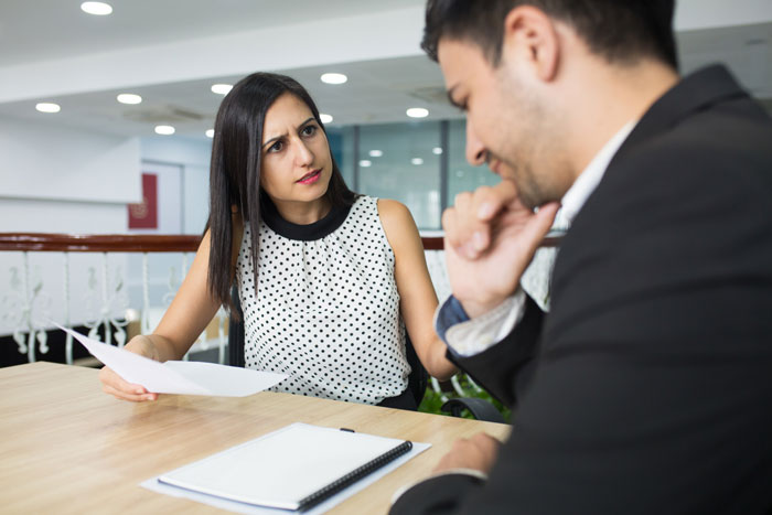 An upset woman holds papers across a table from a man; an office food thief likely facing firing. An upset woman holds papers across a table from a man; an office food thief likely facing firing.