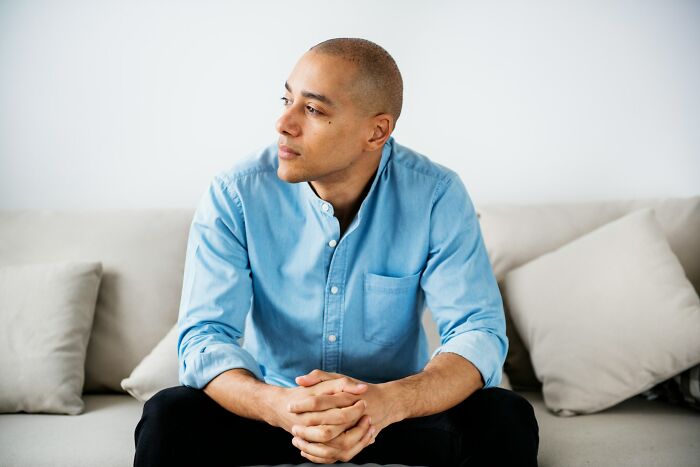 A man with shaved head, wearing a light blue shirt, sitting on a couch with clasped hands, looking off to the side, reflecting on why men are suffering.
