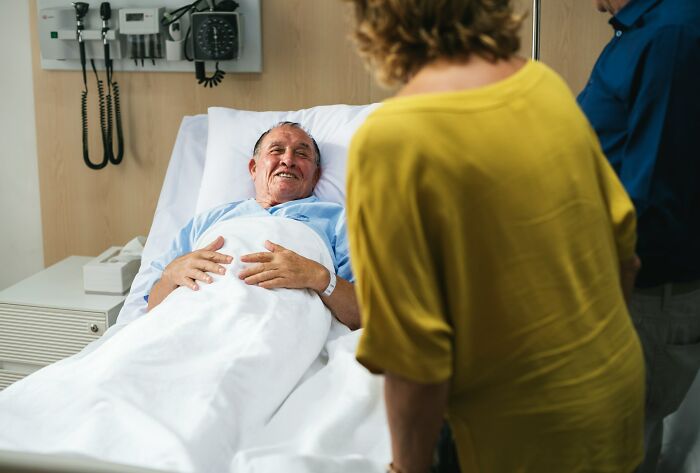 A smiling elderly man in a hospital bed with a woman in a yellow shirt visiting him, reflecting on deeply immoral things.