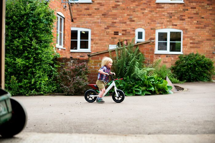 Child riding a balance bike outside a brick house with surrounding greenery