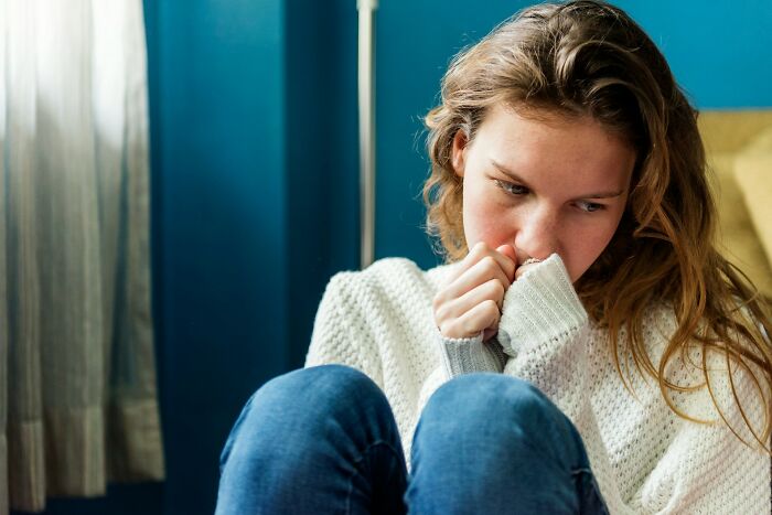 A young woman with long hair in a white sweater and blue jeans, looking down with a worried expression, feeling cringey.