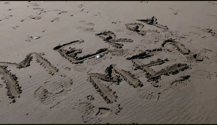 The word 'MERCURIAL' carved into the sand on a beach, illustrating humorous engagement stories and love's beautiful chaos.