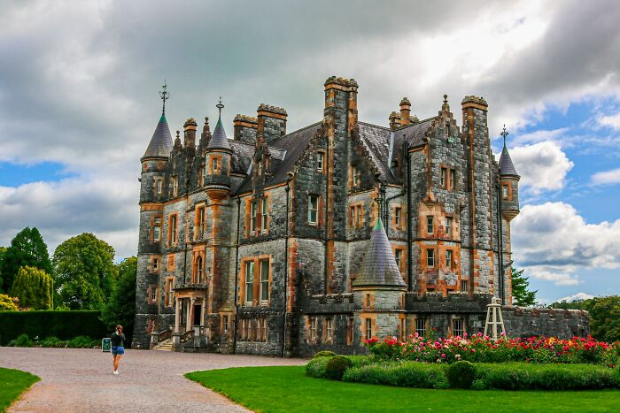 A grand, historic castle with intricate stonework, turrets, and lush gardens under a cloudy sky. Reminiscent of a fairy tale engagement story.