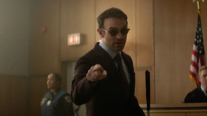 Charlie C*x como Demolidor: Nascido de Novo, usando óculos escuros e terno, gesticulando em um tribunal.