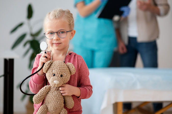 A young girl, wearing glasses and holding a stethoscope to a teddy bear, while her Grandkid's Allergies are being discussed by adults.