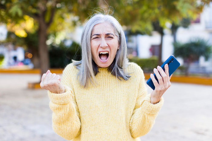 A woman with gray hair and a yellow sweater screams while holding a phone, reacting to her grandkids allergies.