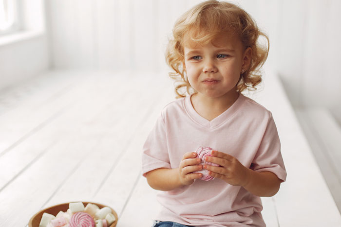A young blonde girl looking distressed, holding a pink marshmallow. She seems to be experiencing allergies.